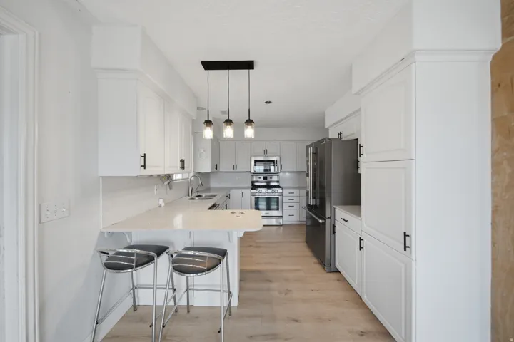 Modern galley kitchen featuring white cabinetry, stainless steel appliances including a double wall oven, quartz countertops, pendant lighting, wood-finish flooring, and a breakfast bar with seating