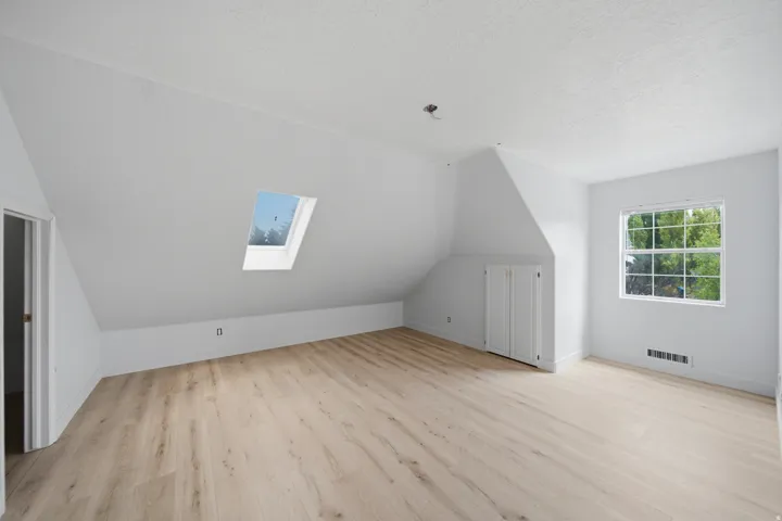 Bright attic room featuring sloped ceilings, light wood-finish flooring, and abundant natural light from a skylight and large multi-pane window