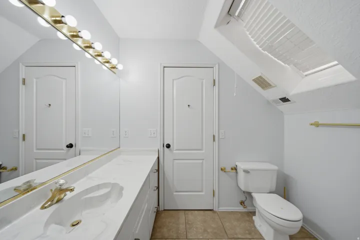 Primary bathroom featuring dual vanities with white cabinetry, brushed brass hardware and fixtures, tile flooring, and natural light from skylights