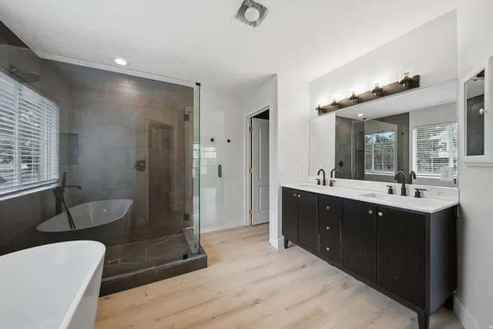 Spa-inspired primary bathroom featuring a freestanding soaking tub, walk-in shower with frameless glass enclosure and tile surround, dual-sink vanity with dark cabinetry and white countertop, wood-finish flooring, and natural light from multiple windows