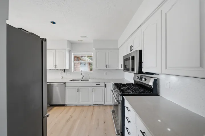 Bright, contemporary kitchen showcasing white shaker-style cabinetry, stainless steel appliances including a dual-oven range, quartz countertops, a tile backsplash, and wood-finish flooring throughout the open layout