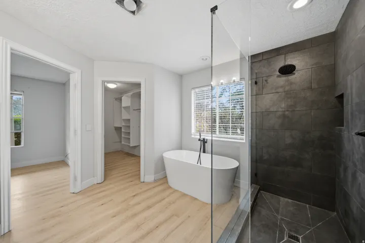 Freestanding soaking tub, walk-in shower with dark tile surround, glass shower enclosure, wood-finish flooring, and built-in closet shelving