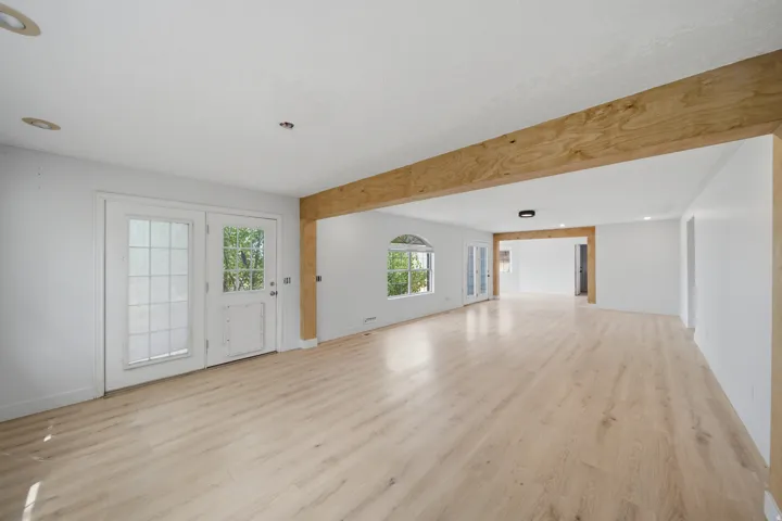 Spacious open-concept living area featuring light wood-finish flooring, exposed timber beam ceiling, and abundant natural light from multiple windows and glass doors