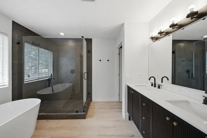 Spa-inspired primary bathroom featuring dual vanities with white quartz countertops and matte black fixtures, a freestanding soaking tub, a walk-in shower with dark tile surround and glass enclosure, wood-finish flooring, and brass-finished lighting fixtu