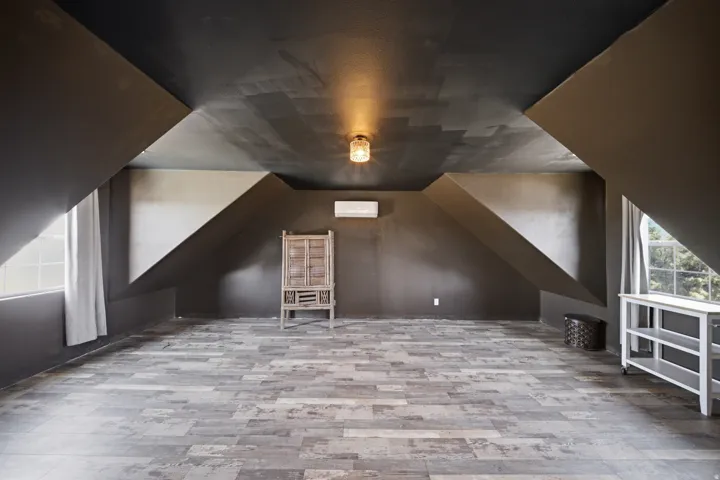 Spacious attic room featuring vaulted ceilings with exposed beams, wood-finish flooring throughout, and abundant natural light from dual windows