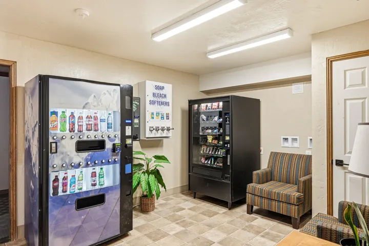 Convenient break room featuring dual vending machines with beverage and snack selections, tile flooring, recessed and linear ceiling lighting, striped upholstered seating, potted greenery, and white six-panel doors providing access to adjacent spaces