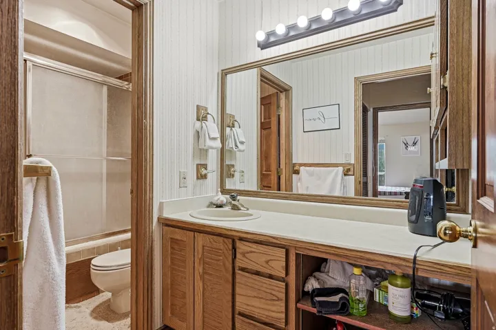 Vanity with integrated sink, wood-finish cabinetry, full-width mirror with overhead lighting, and an adjacent shower-tub combination