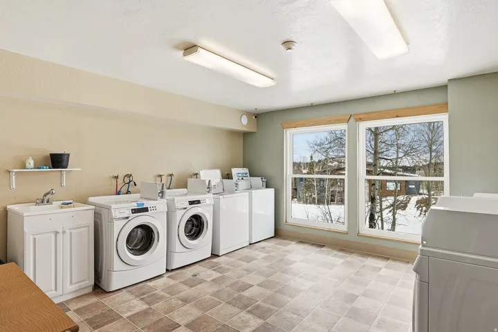 Spacious laundry room featuring dual front-load washers and dryers, extensive white cabinetry with upper shelving, tile flooring, and abundant natural light from large glass doors overlooking the snow-covered landscape beyond