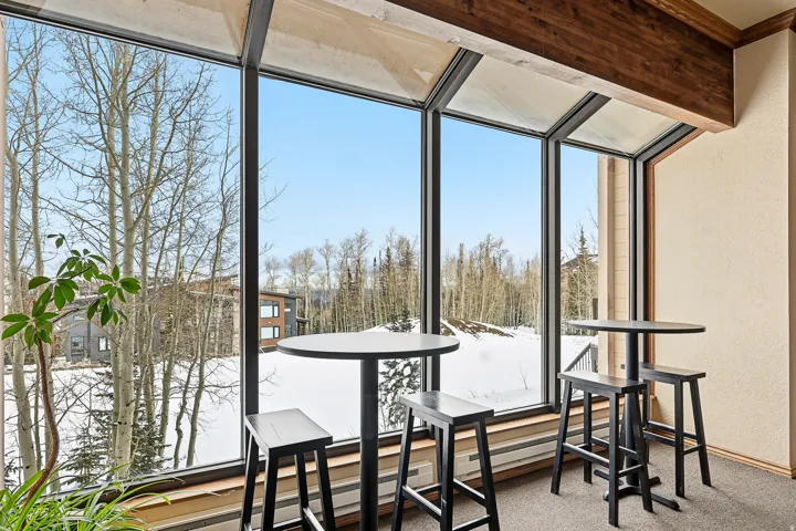 Modern sunroom with expansive floor-to-ceiling windows framing forested landscape and seasonal vistas, wood-beam ceiling detail, high-top bar seating with black metal stools, and polished concrete flooring throughout