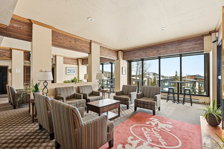 Sophisticated lounge area showcasing exposed wood beams, floor-to-ceiling windows with natural light, striped upholstered seating arrangements, wood-finish flooring with geometric patterned accent rug, and direct access to an expansive outdoor terrace wit