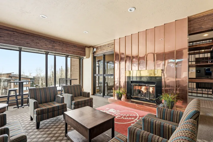 Sophisticated penthouse living room showcasing floor-to-ceiling windows with cityscape exposure, copper-tone accent wall paneling, built-in shelving with dark wood finishes, recessed ceiling lighting, and a gas fireplace with decorative surround