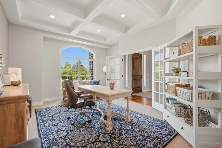 Bright home office featuring coffered ceilings, large arched windows framing natural light, wide-plank wood-finish flooring, and built-in white cabinetry with open shelving