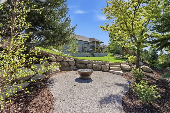 Backyard retreat featuring a circular fire pit surrounded by stacked stone seating walls, gravel base, and mature landscaping with mixed deciduous trees