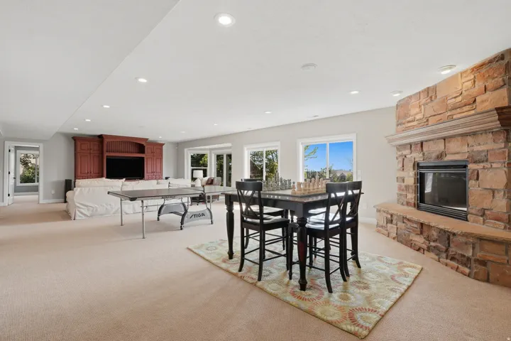 Expansive recreation room featuring soaring ceilings with recessed lighting, a wood-burning fireplace with brick surround, black dining table with seating for four, billiards table, built-in cabinetry in rich burgundy tones, and abundant natural light fro