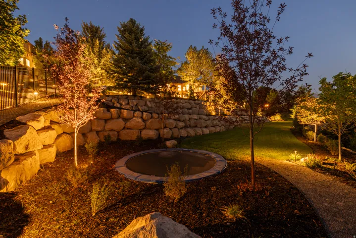 Expansive backyard featuring tiered stone retaining walls, manicured lawn, gravel pathways, and mature trees with integrated landscape lighting