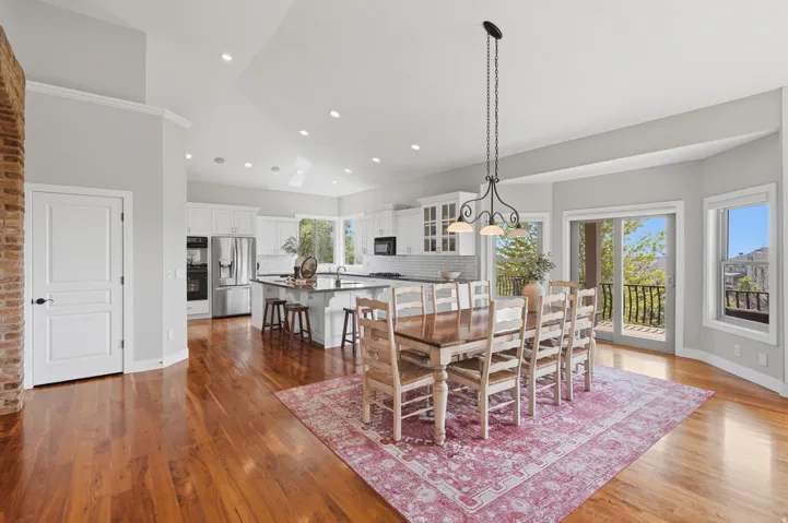 Open-concept living space featuring hardwood flooring throughout, a large dining area with seating for eight, and a modern kitchen with stainless steel appliances and white cabinetry