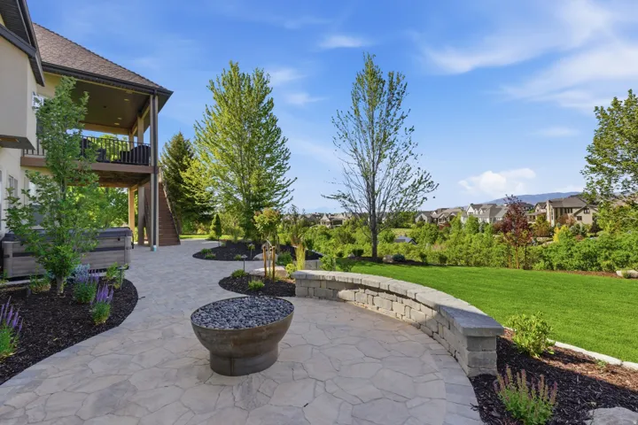 Expansive patio featuring natural stone hardscaping with curved retaining walls, mature landscaping with mixed evergreen and deciduous trees, manicured lawn, and integrated planter beds with seasonal plantings