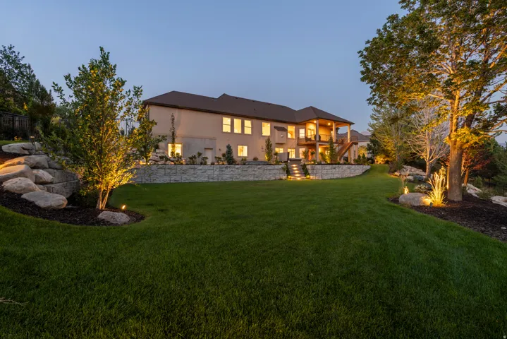 Expansive manicured lawn anchored by mature landscaping and uplighting, with a two-story stone-and-stucco residence featuring illuminated multi-pane windows, professional exterior lighting design, and tiered hardscaping throughout the grounds