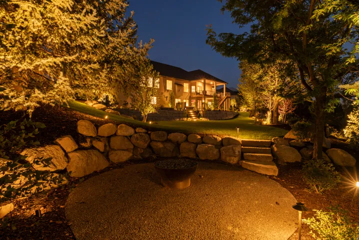 Elegant home illuminated by professional landscape lighting, featuring mature trees framing the property and a curved stone seating wall anchoring the grounds