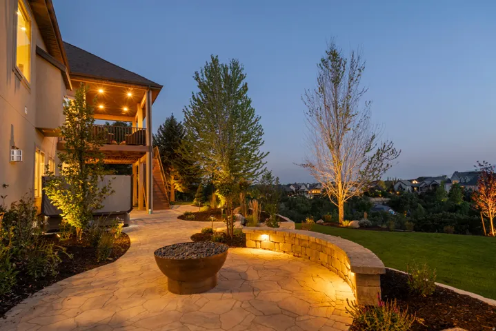 Elegant outdoor entertaining space featuring a curved stone patio with ambient landscape lighting, mature specimen trees, and a built-in fire pit