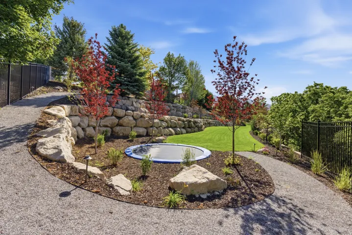Expertly landscaped backyard oasis featuring a circular fire pit with stone seating, mature specimen trees with seasonal foliage in red and burgundy tones, lush lawn, decorative boulder accents, gravel pathways, and black metal perimeter fencing for defin