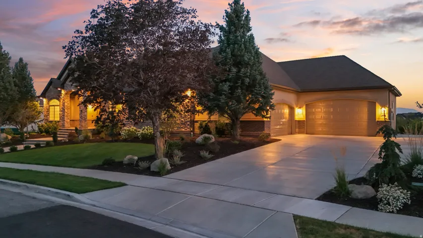Elegant home showcasing warm architectural lighting, a circular driveway with wide concrete apron, mature landscaping with specimen trees, manicured lawn, and a three-car garage with dark roofing and stone/brick exterior finishes