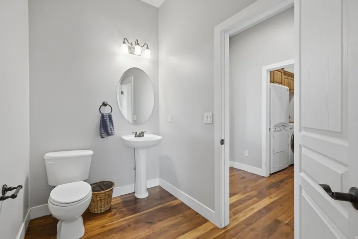 Powder room featuring a pedestal sink with chrome faucet, oval mirror with integrated lighting, ceramic toilet, and warm wood-finish flooring throughout