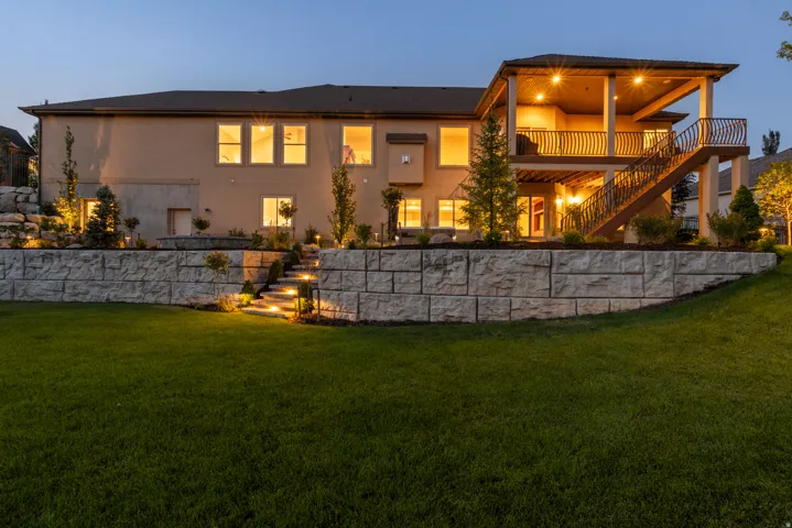 Modern two-story residence with illuminated interior spaces, featuring a composite stone retaining wall, manicured lawn, mature landscaping with specimen trees, a covered upper-level deck with railings, and exterior accent lighting throughout the grounds