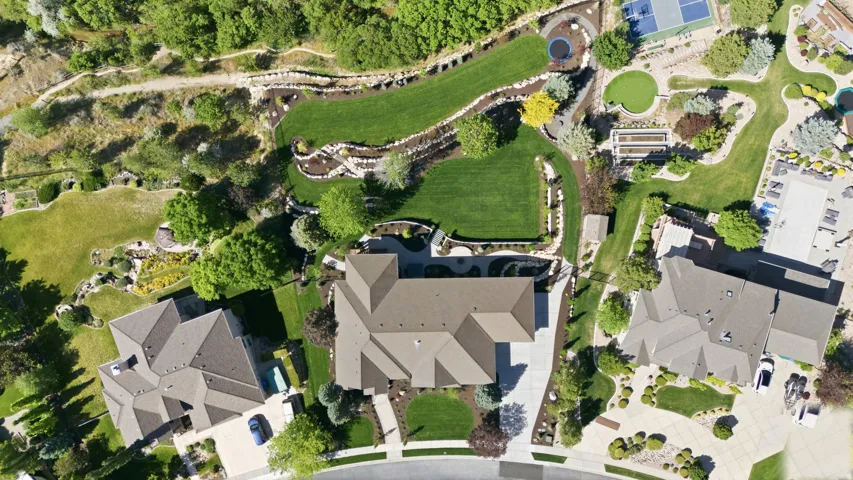 Aerial view showcasing a substantial two-story residence with multiple roof peaks and architectural depth, situated on expansive grounds featuring manicured lawns, mature landscaping, and integrated hardscaping elements