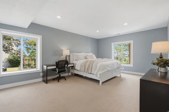 Bright, light-filled primary bedroom suite featuring expansive windows framing natural landscape views, neutral carpet flooring, soft recessed ceiling lighting, and generous square footage ideal for a full bedroom layout with dedicated workspace and dress