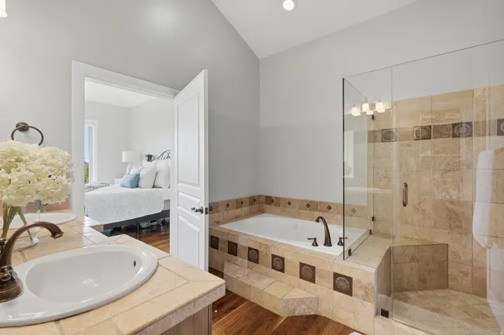 Spa-inspired primary bathroom featuring a soaking tub with decorative tile surround, a walk-in shower with glass enclosure and travertine-look stone finishes, dual vanity with wood-finish countertop and round vessel sinks, and warm lighting