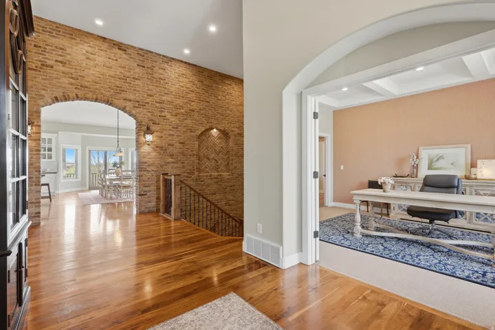 Elegant foyer showcasing rich wood-finish flooring, exposed brick accent wall with arched architectural detailing, recessed ceiling lighting, and a curved archway entry leading to adjoining spaces