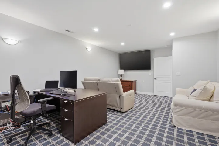 Versatile basement recreation space featuring a geometric patterned area rug, recessed ceiling lighting, wall-mounted flat-screen television, dark wood desk with ergonomic seating, upholstered seating arrangements, and white paneled doors providing access