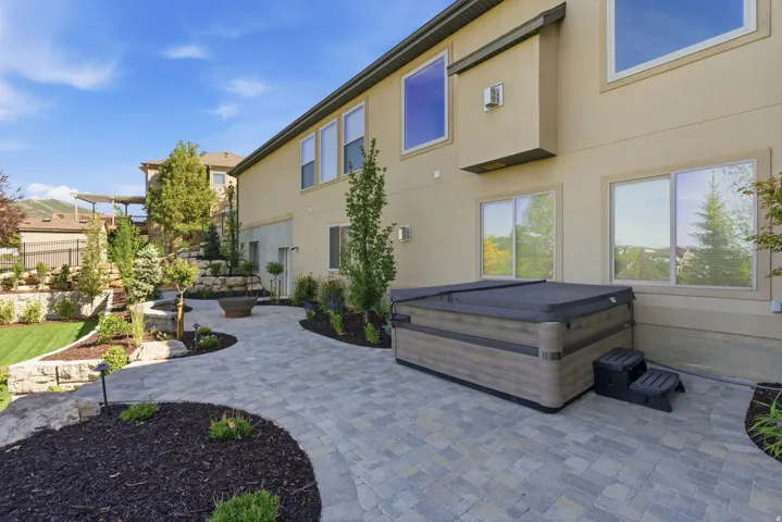 Expansive paver patio with integrated hot tub and lounge seating area, anchored by mature landscaping with ornamental trees and mulched garden beds