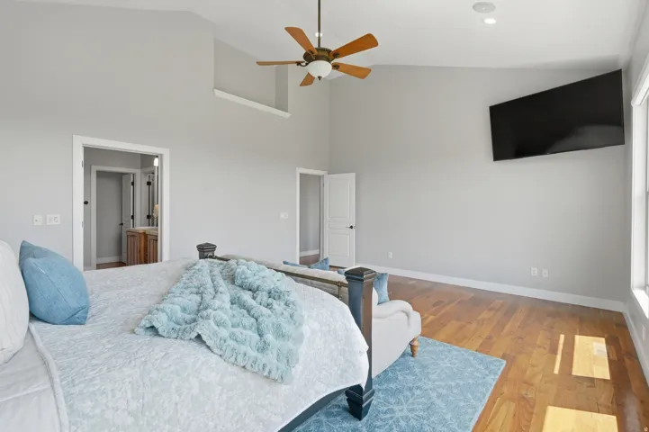 Spacious primary bedroom with wood-finish flooring, ceiling fan with integrated lighting, wall-mounted flat-screen television, and dual doorway access to adjoining spaces