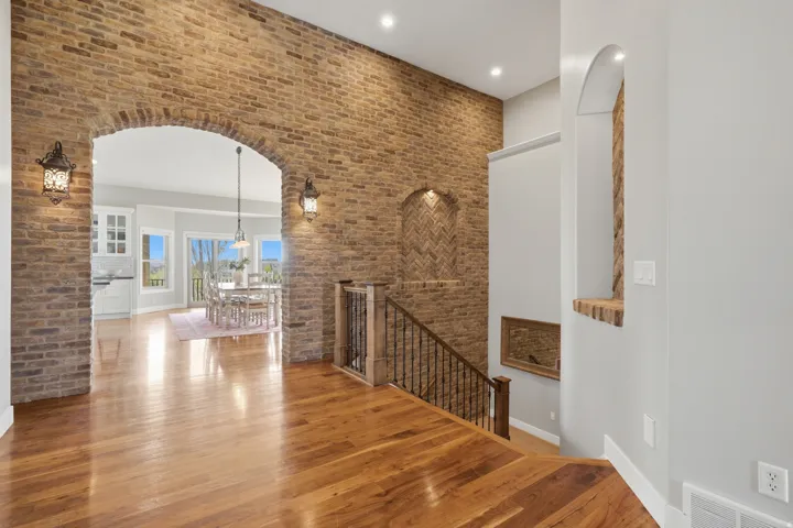 Elegant foyer showcasing exposed brick archway with arched opening, hardwood flooring, wrought iron stair railing, recessed lighting, and sightlines to bright interior spaces with natural light