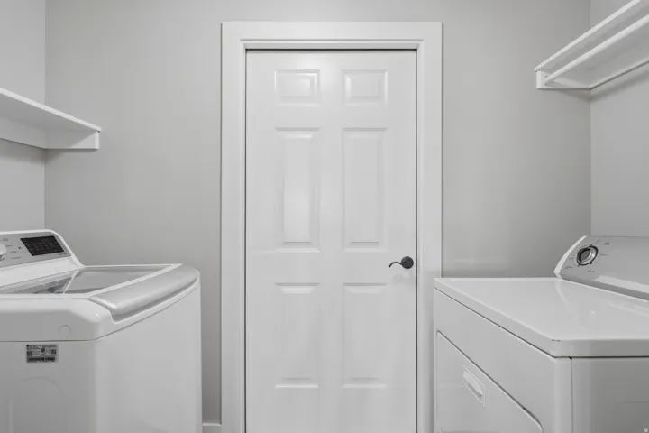Efficient laundry room featuring side-by-side washer and dryer appliances, white six-panel entry door, bright recessed lighting, and clean white cabinetry with ample storage shelving