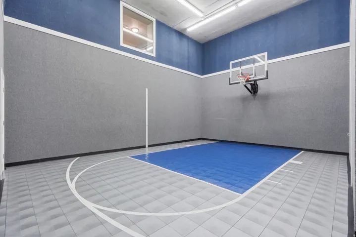Indoor recreational court featuring a regulation basketball hoop with backboard, polished tile flooring with court markings, blue synthetic playing surface, and acoustic ceiling with integrated lighting