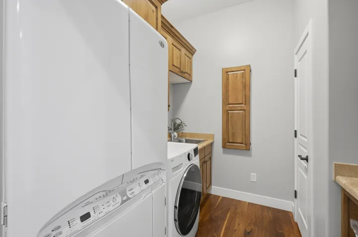 Dedicated laundry room featuring stacked front-load washer and dryer with white cabinetry, wood-finish upper storage, wood-finish flooring, and abundant natural light from recessed ceiling fixtures