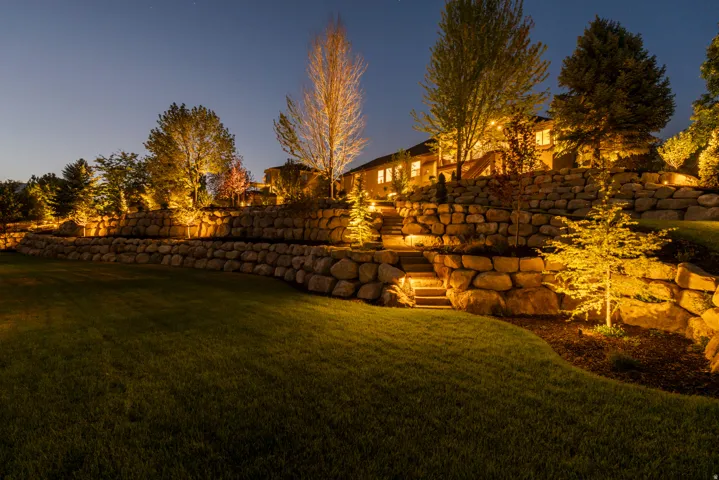 Expertly landscaped grounds featuring a manicured lawn, mature specimen trees with golden uplighting, and multi-tiered stone retaining walls