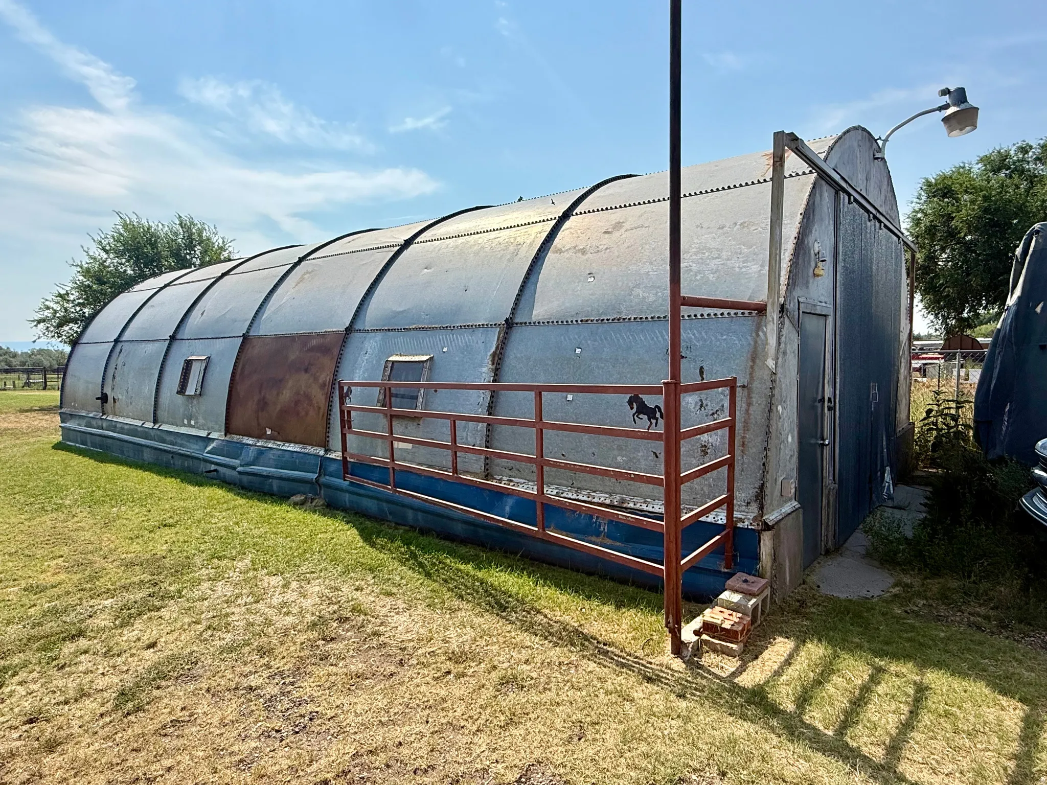 Corrugated metal Quonset hut structure with an arched roof, featuring a side entry door and a metal fence