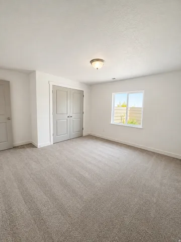 Spacious bedroom with neutral carpet flooring, abundant natural light from a large double-hung window, built-in double-door closet with paneled doors, ceiling-mounted light fixture, and clean white walls throughout