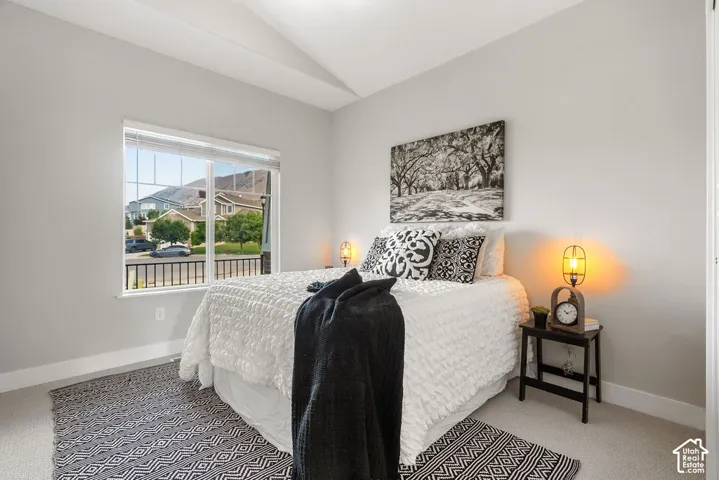Bright primary bedroom with expansive windows framing mountain vistas, neutral wall finishes, geometric patterned area rug, bedside tables with integrated lighting, and a black accent piece at the foot of the bed