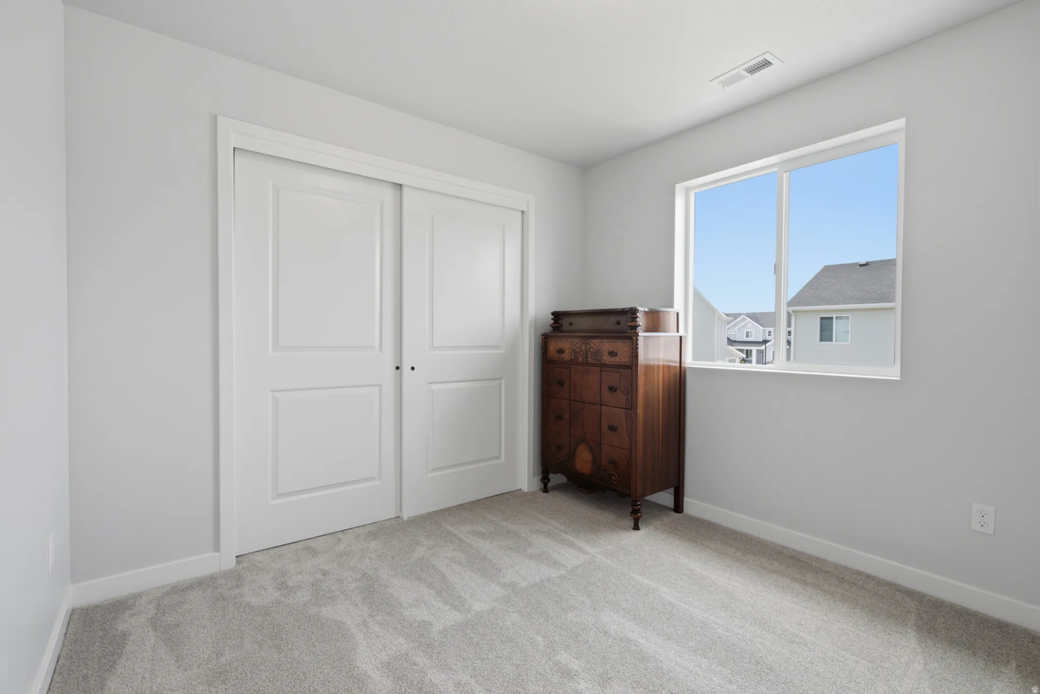 Bright secondary bedroom featuring dual closets with white panel doors, carpet flooring, and abundant natural light from a large window overlooking the neighborhood
