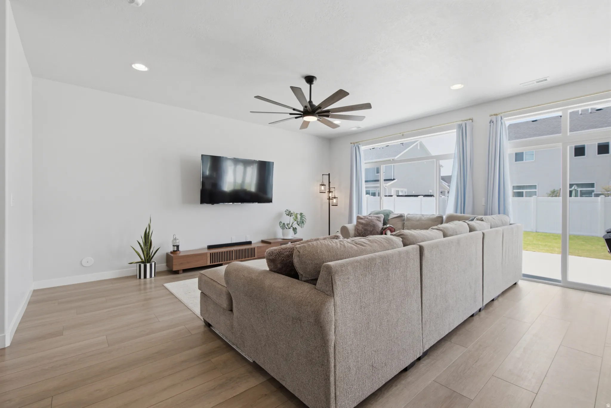 Modern living room with expansive wood-finish flooring, abundant natural light from sliding glass doors and large windows, ceiling fan with bronze finish, wall-mounted flat-screen television, and recessed lighting throughout