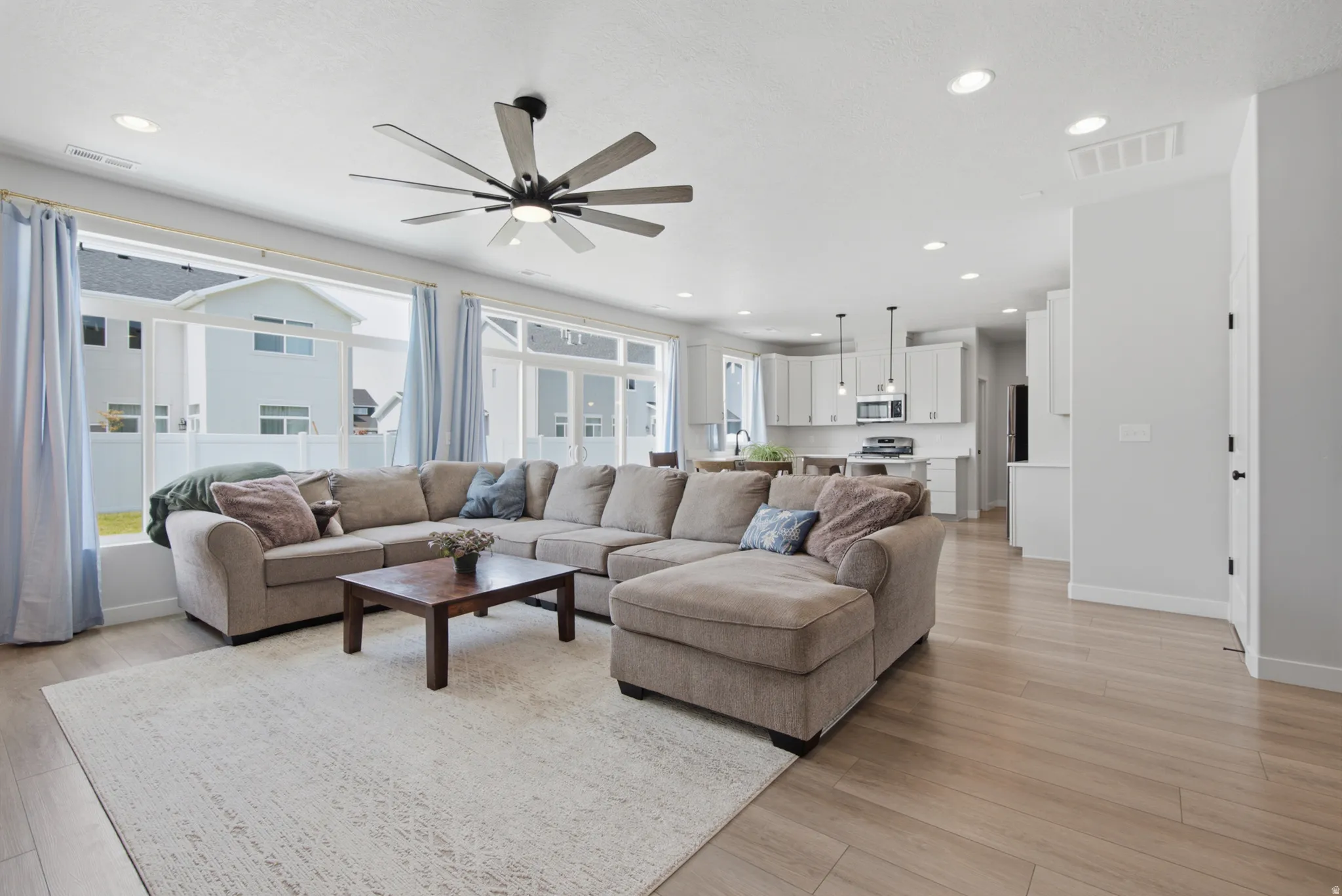 Spacious open-concept living area showcasing wood-finish flooring, a large sectional configuration with chaise, modern ceiling fan with brushed metal finish, recessed lighting throughout, and abundant natural light from expansive sliding glass doors and w