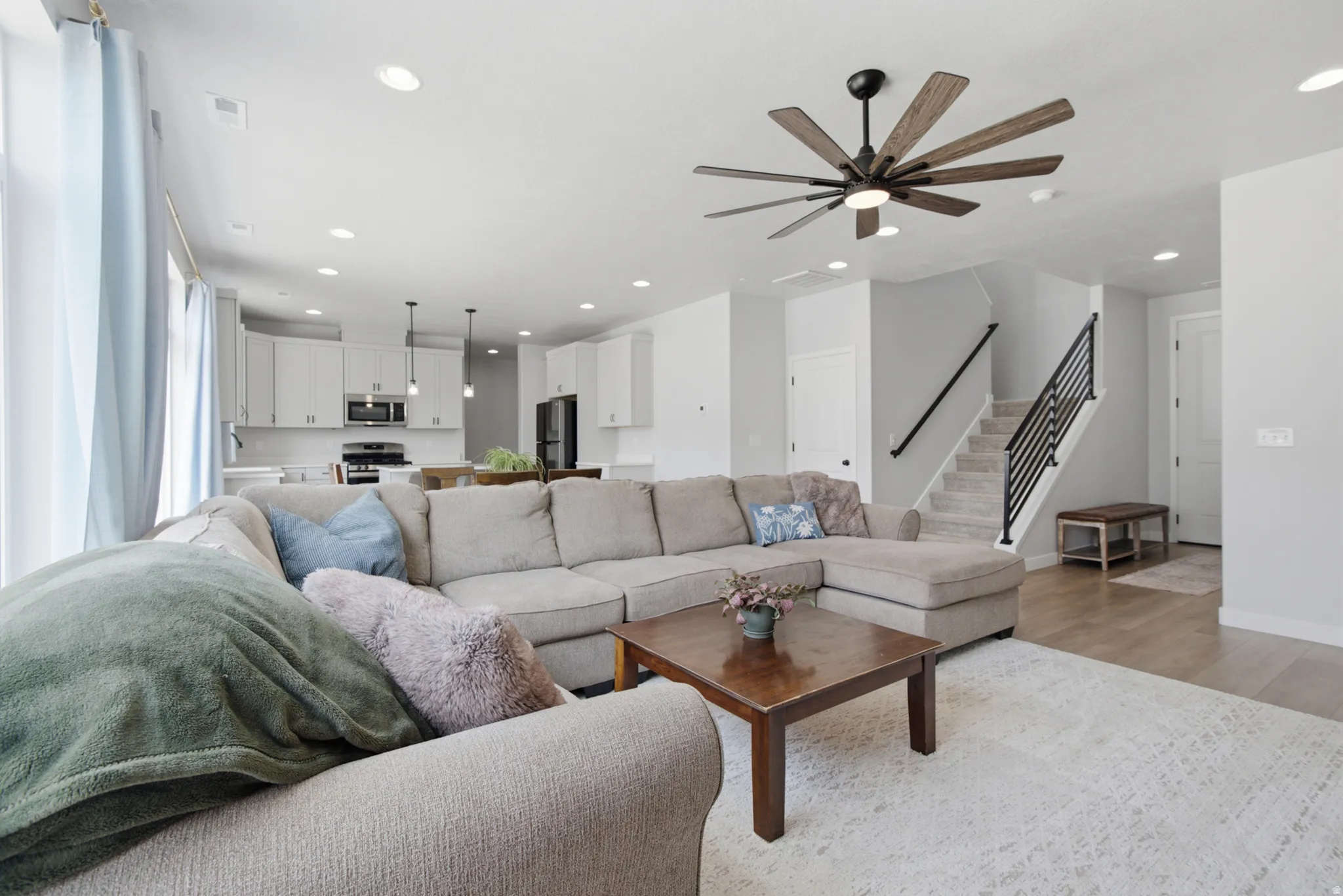 Open-concept living area featuring a sectional-ready layout with recessed ceiling lights, a modern ceiling fan with wood-finish blades, and direct sightlines to the kitchen