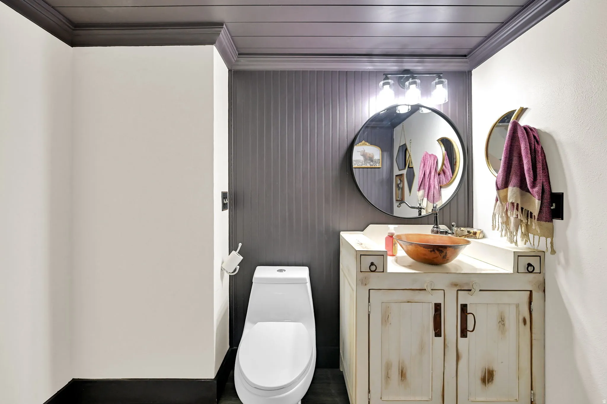 Modern bathroom featuring a sleek one-piece toilet, vanity with dual cabinet storage and wood-finish detailing, ceramic vessel sink, polished countertop, circular wall-mounted mirror with integrated lighting, vertical shiplap accent wall, dark ceiling wit