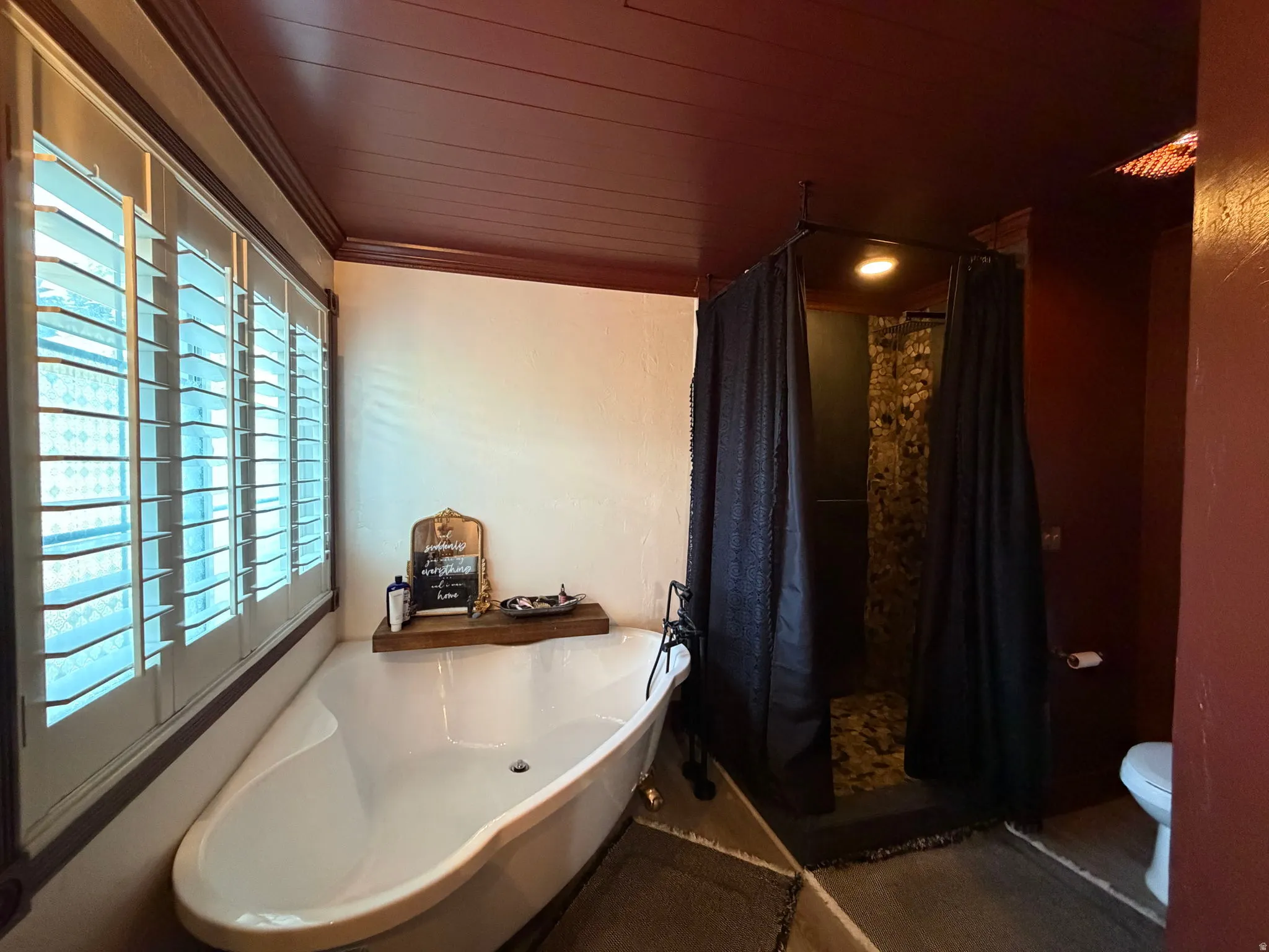 Freestanding bathtub with dark bronze fixtures, plantation shutters, and a walk-in shower with a dark curtain