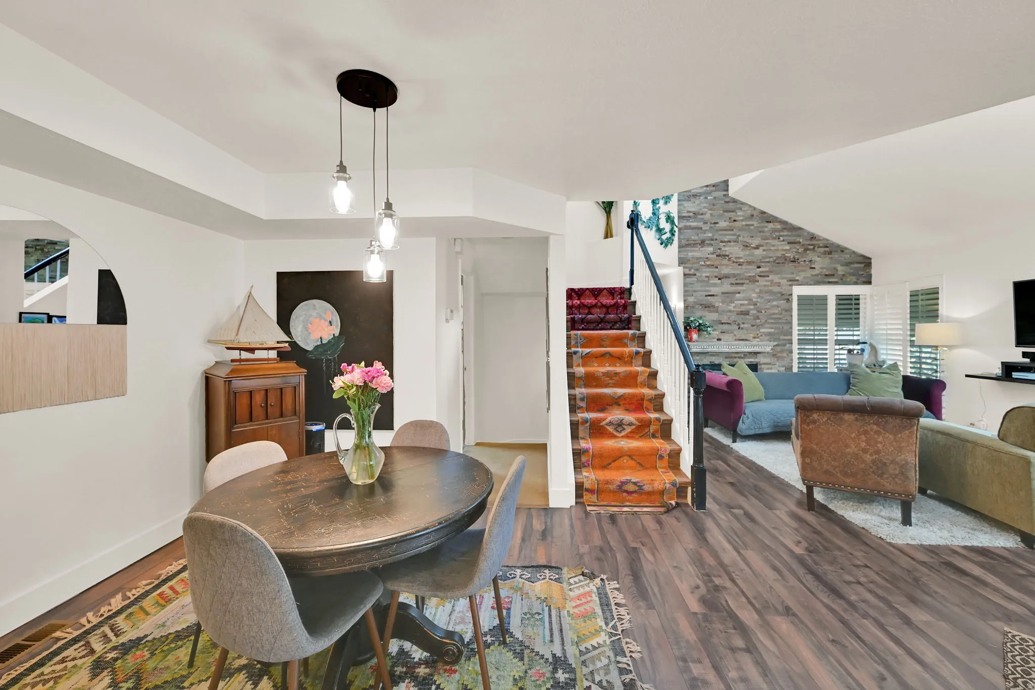 Wood-finish flooring throughout, a dining area with a multi-light pendant fixture, staircase with decorative runner, a stone accent wall, and interior window shutters